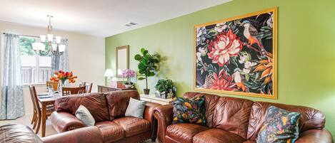 A living room with a brown leather sofa, a dining table with chairs, and a large floral painting on the wall.