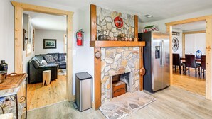 A living room with a stone fireplace, a black leather sofa, a wooden coffee table, and a modern kitchen with a stainless steel refrigerator. - Deck & Scenic View: Historic Cabin in Piney Creek! (Piney Creek)