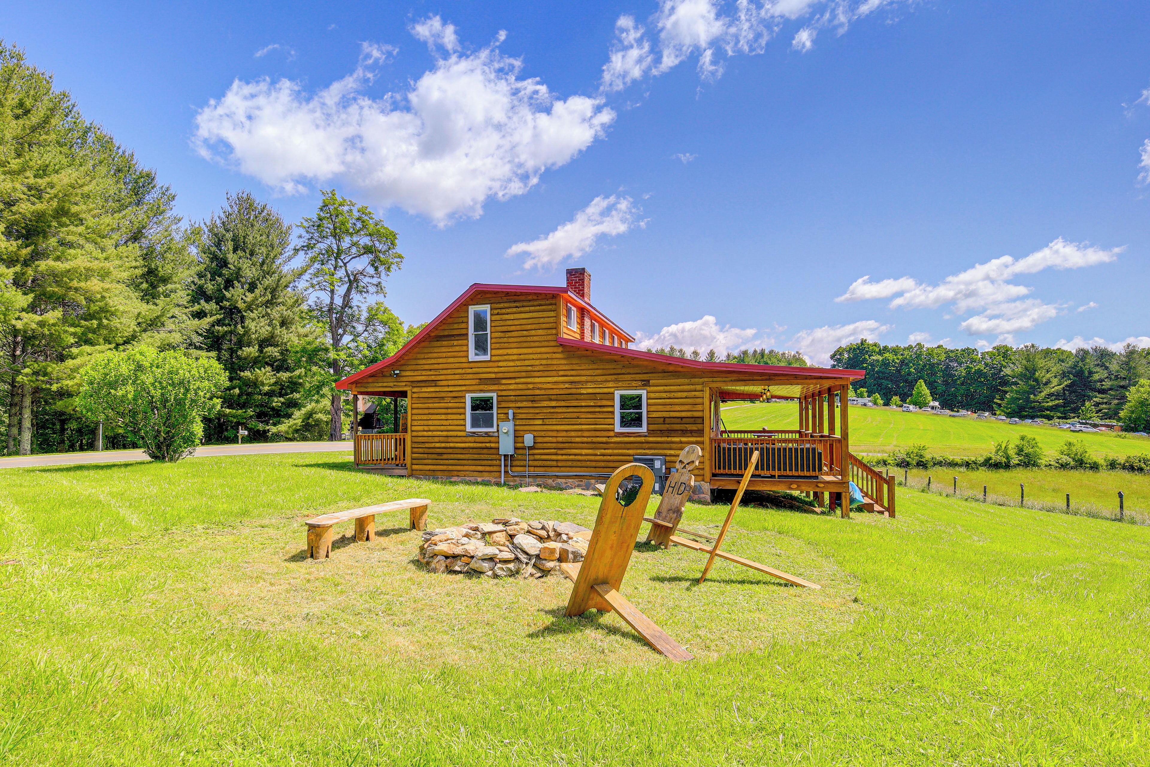 A wooden cabin with a red roof, a porch, and a stone fire pit in a grassy field.