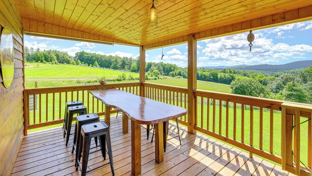 A wooden deck with a table and stools, overlooking a green landscape. - Deck & Scenic View: Historic Cabin in Piney Creek! (Piney Creek)