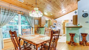 A wooden dining table with chairs, a chandelier, and a view of the outdoors through large windows. - Serene Maggie Valley Retreat w/ Deck & Hot Tub! (Maggie Valley)