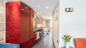 A modern kitchen with red cabinetry, a stone accent wall, and a view to the living area. - Architecturally designed fishing warehouse conversion (Newlyn)
