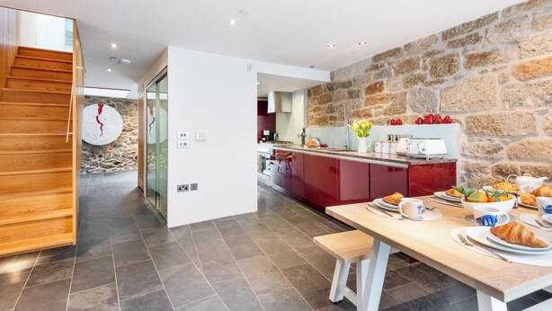 A modern kitchen with a stone wall, wooden staircase, and a breakfast setup with croissants and orange juice. - Architecturally designed fishing warehouse conversion (Newlyn)