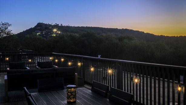 Eine Holzterrasse mit Sitzgelegenheiten und Lichterketten, mit Blick auf einen Hügel mit Gebäuden.
