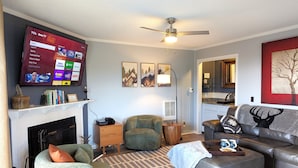 Ein Wohnzimmer mit Kamin, einem Wand-Fernseher und Deckenventilator, mit einer Couch und einem Kissen mit Hirschmotiv.