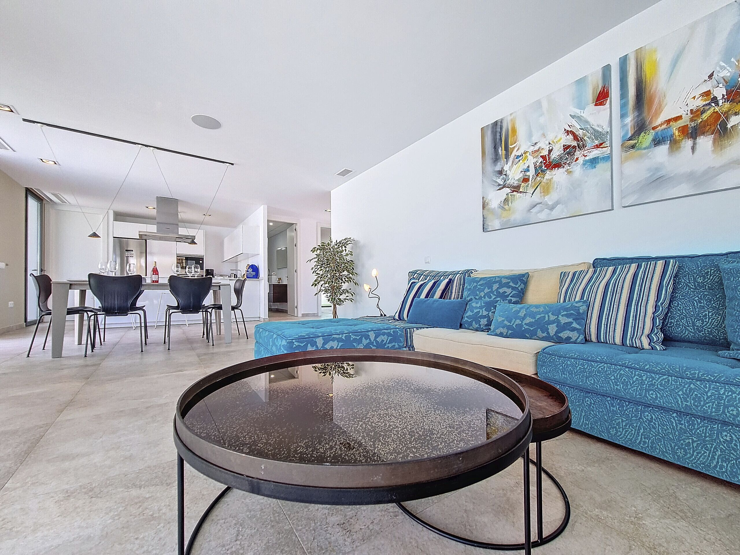 A modern living room with a blue sofa, a round coffee table, and a dining area with chairs and a table.