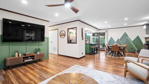 A spacious living area with a wooden floor, a ceiling fan, a flat-screen TV, a dining area with a table and chairs, and a kitchen visible through an open door. - Rolling Hills/Hot Tub/Firepit/Mins to ACE+NRG (Oak Hill)