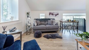 Un salon moderne avec un canapé gris, une table à manger avec des chaises, un luminaire suspendu et une vue sur l’extérieur grâce à de grandes fenêtres.