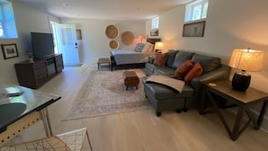 A modern living room with a sofa, coffee table, and TV. There is a dining area with a table and chairs. The room has a large window and a door leading outside.