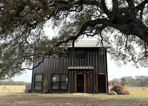 A two-story building with a dark exterior, large windows, and a door with a yellow frame. - Private, cabin with endless Hill Country views. Horse & dog friendly! (Pipe Creek)