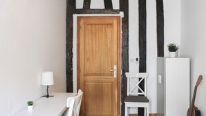 A room with a wooden door, a white table with a lamp, a white chair, a small stool, a white cabinet, and a guitar leaning against the wall.