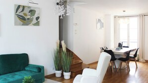 A modern living room with a green sofa, a glass coffee table, and a white armchair. There is a dining area with a table and chairs, and a staircase leading to another level.