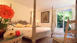 Een hemelbed met een witte spreidek, een nachtkastje met een bloevas en een stoel met een kussen.