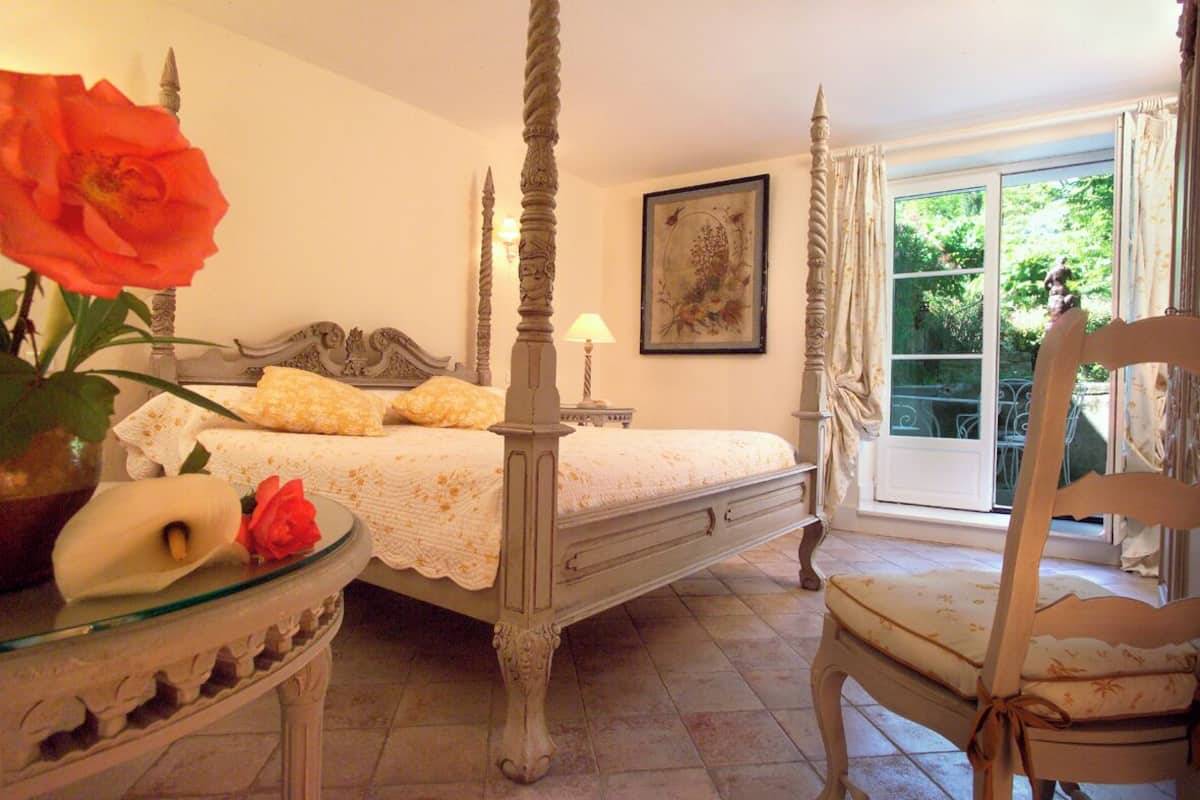 A four-poster bed with a white bedspread, a bedside table with a vase of flowers, and a chair with a cushion.