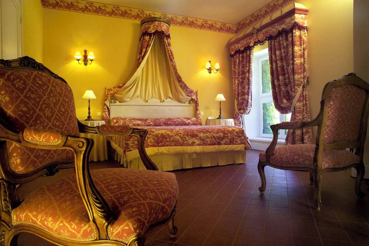 A bedroom with a four-poster bed, ornate chairs, and a large window with curtains.