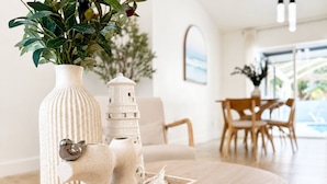 A modern interior with a wooden table displaying decorative items, a white chair, and a dining area with wooden furniture. - House w/ Pool, Hot Tub & Backyard (Cape Coral)