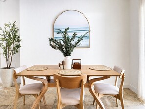 A dining area with a wooden table, four chairs, a potted plant, a framed picture, and a vase with greenery. - House w/ Pool, Hot Tub & Backyard (Cape Coral)