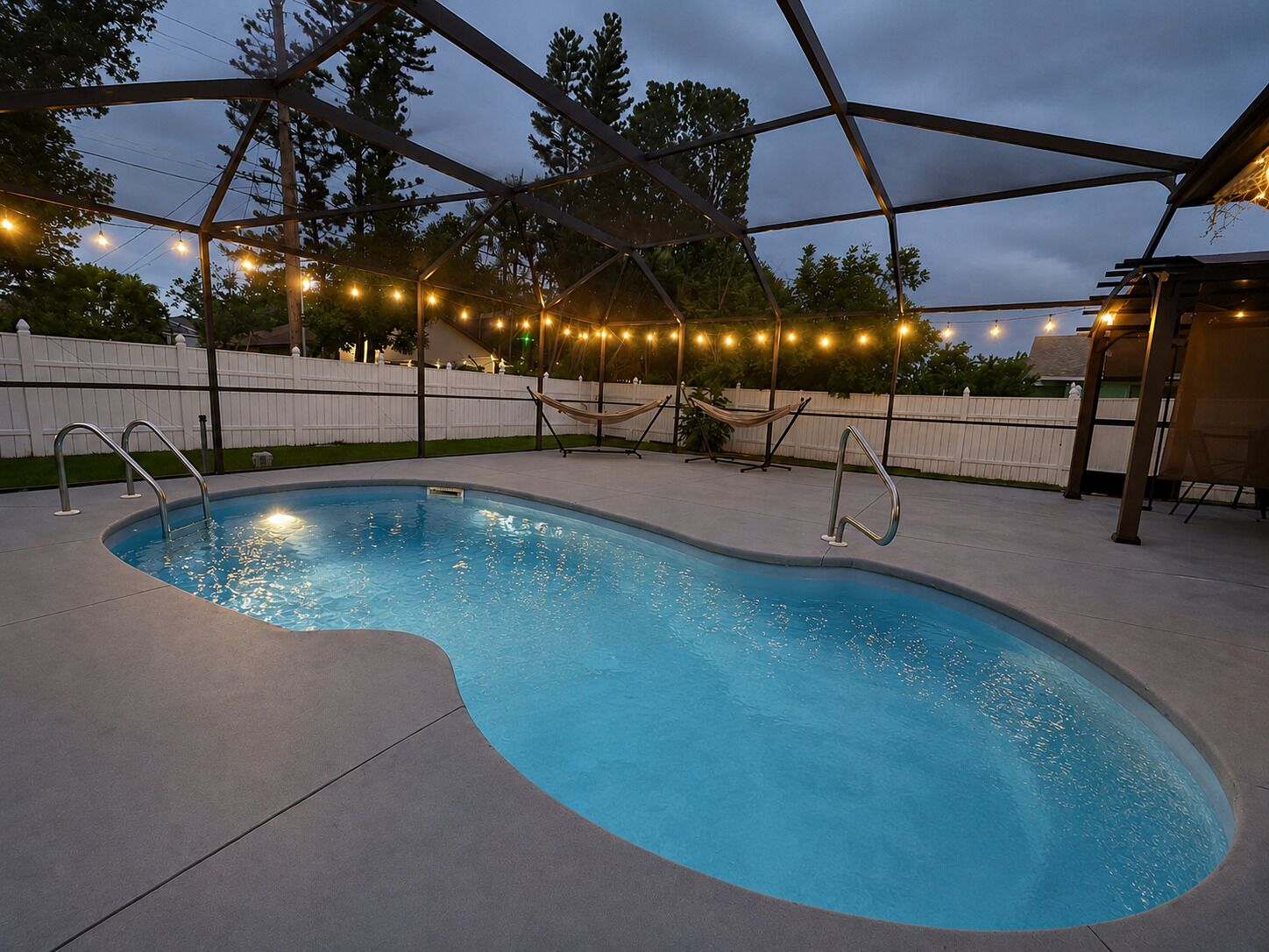 Una piscina con estructura metálica y barandillas, rodeada por una cerca blanca y árboles.