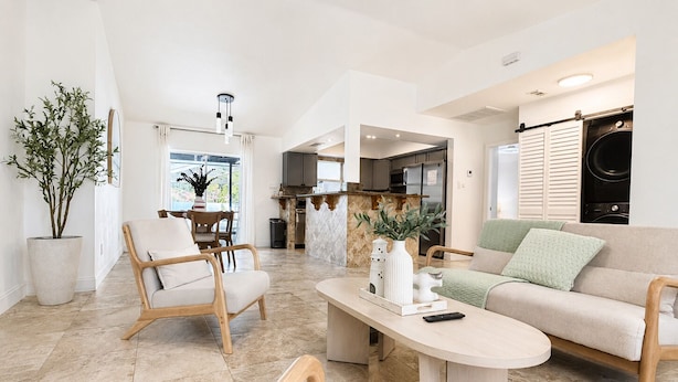 A modern living room with a sofa, chairs, a coffee table, and a kitchen in the background. - House w/ Pool, Hot Tub & Backyard (Cape Coral)