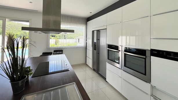 Een moderne keuken met witte kasten, roestvrijstalen apparatuur, een groot raam met jaloezieën, en een eetgedeelte met een tafel en stoelen.