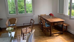 Un coin repas avec une table et des chaises en bois, un vase rempli de fleurs et une vue sur la verdure grâce à de grandes fenêtres.
