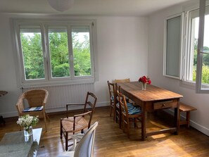 Een eetgedeelte met een houten tafel en stoelen, een vaas met bloemen en uitzicht op groen door grote ramen.