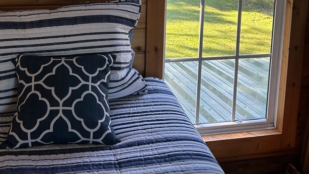 A wooden cabin room with a bed, a framed certificate on the wall, and a large window overlooking a grassy area. - Enjoyable 2-bedroom bungalow with AC close to charming "Beaufort by the Sea" . (Havelock)