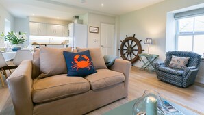 Ein Wohnzimmer mit einem beigefarbenen Sofa, einem gemusterten Sessel, einem hölzernen Steuerrad und einer Küche im Hintergrund.