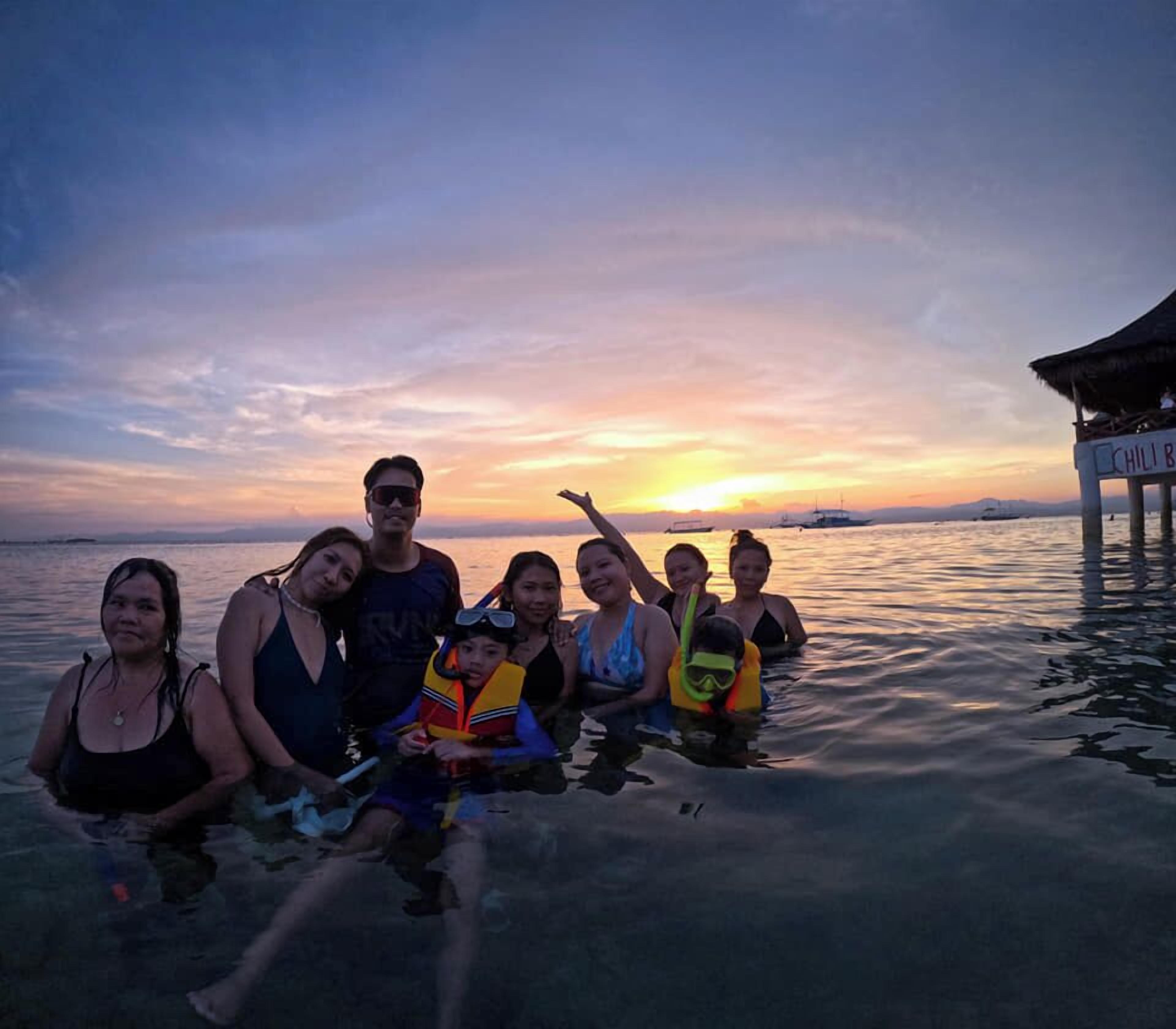 Un groupe de personnes se trouve dans un cadre tropical, avec un coucher de soleil en arrière-plan.