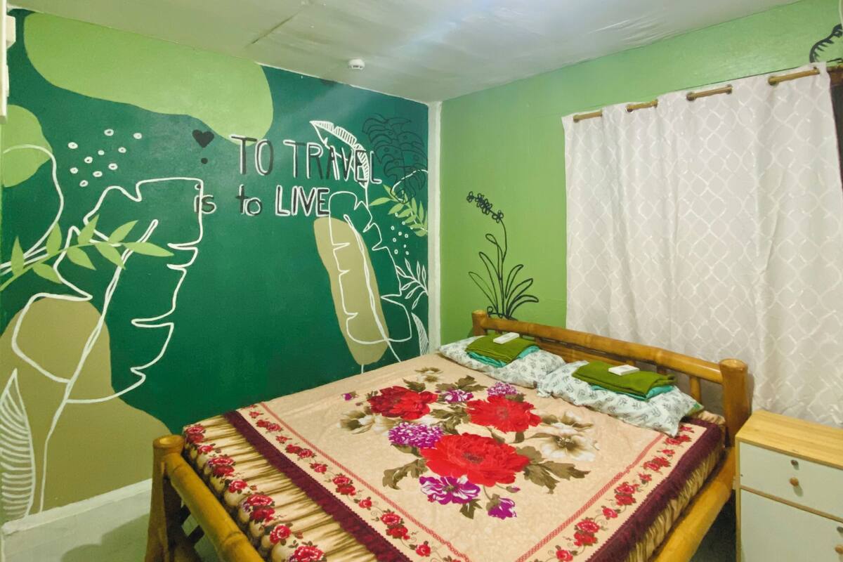 Ein Schlafzimmer mit einem Bett, einer grünen Wand mit Wandbild zum Thema Reisen, einem Holz-Kopfteil, einem weißen Vorhang und einem kleinen Nachttisch.