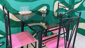 Een eetruimte met een glazen tafel, zwarte metalen stoelen met roze kussens, en een muur met abstracte groene en witte patronen.