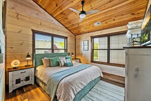 A bedroom with a wooden ceiling, a bed with a green headboard, two bedside tables with lamps, and a large window.