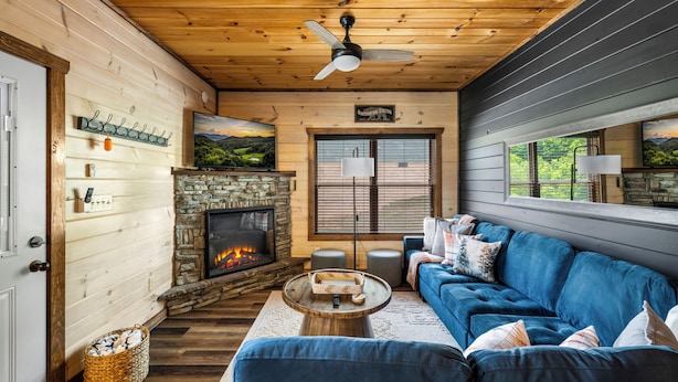 Ein gemütliches Blockhauskaminzimmer mit Kamin, Holzwänden, einem blauen Sofa und einem Deckenventilator.