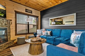 Un salon chaleureux avec une cheminée en pierre, un canapé bleu, une table basse ronde en bois et une grande fenêtre avec des stores.