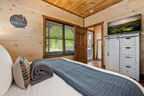 A wooden cabin bedroom with a bed, a large window, a wardrobe, and a painting on the wall.
