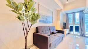 A modern living room with a grey sofa, a large potted plant, a flat-screen TV, and a large window with curtains. - Uptown Parksuites 2 (Taguig)