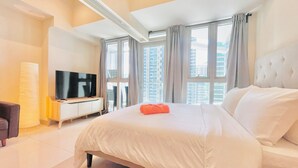 A modern hotel room with a large bed, a flat-screen TV, and a city view through large windows. - Uptown Parksuites 2 (Taguig)