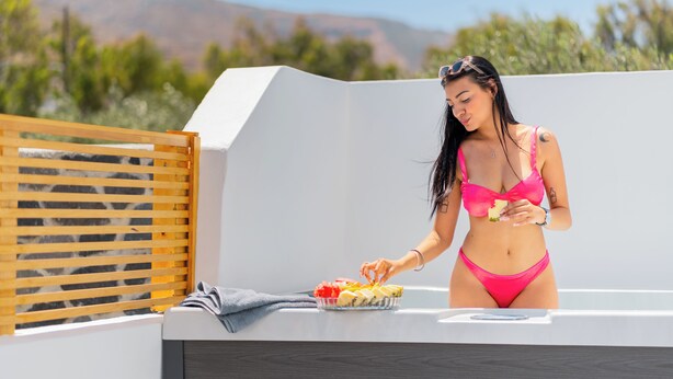 Eine Person in einem rosafarbenen Bikini auf einer Dachterrasse mit einem Teller Essen, einem Holzzaun und Blick auf Berge im Hintergrund.