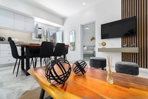 A modern kitchen with white cabinets, a dining area with a wooden table and black chairs, and a flat-screen TV mounted on the wall.