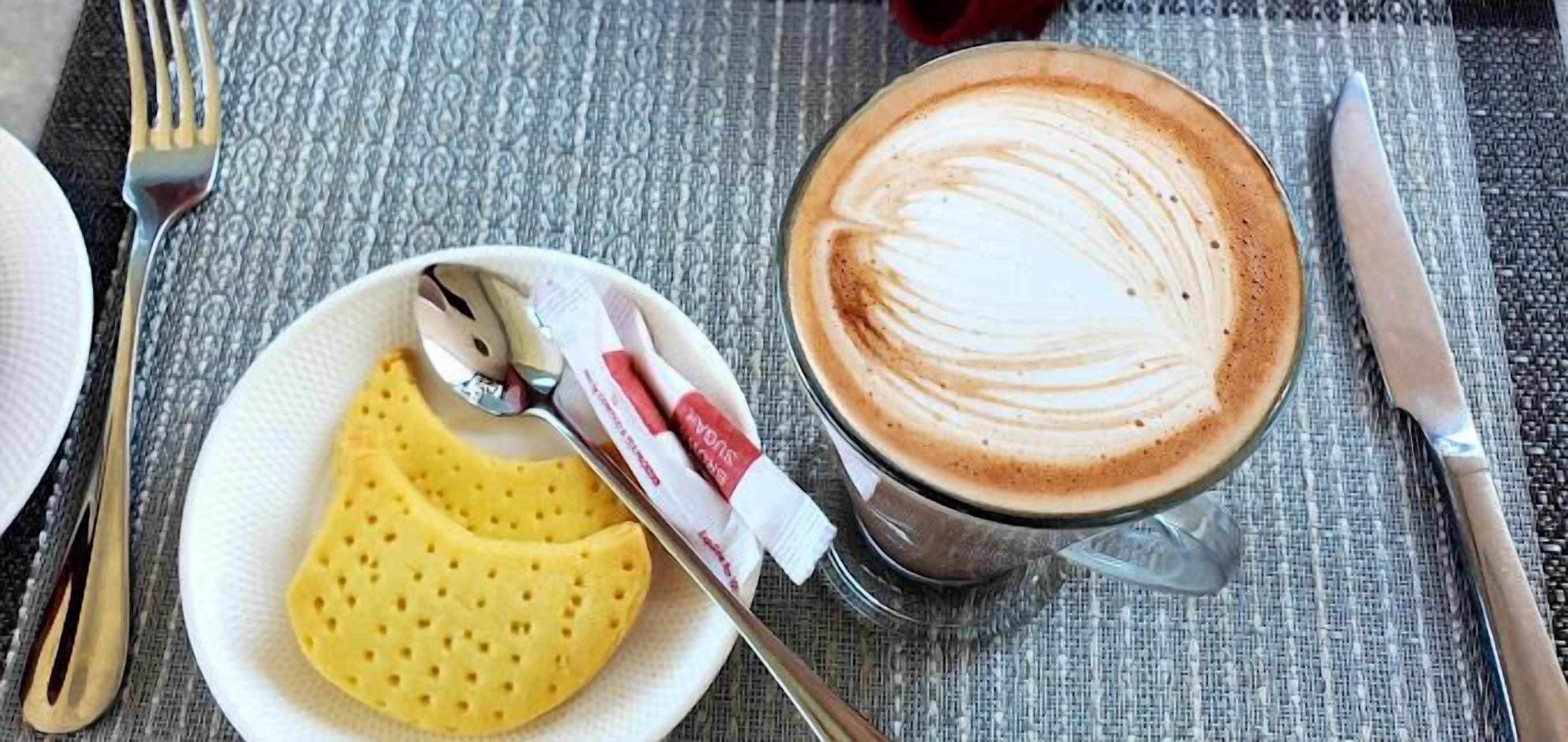 Een kop koffie met latte-art op een geribbelde tafelkleed, een bord met een geel gebakje en bestek.