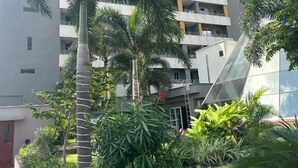 Un edificio de varios pisos con balcones, rodeado de diversas plantas tropicales y un cielo despejado.