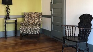 A room with wooden flooring, a patterned armchair, a dark wooden chair, a lamp on a small table, and a sliding door. - Private Downtown Apartment in Red Cloud, in the heart of Willa Cather country (Red Cloud)