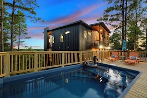A modern cabin with a hot tub and deck chairs, surrounded by trees.