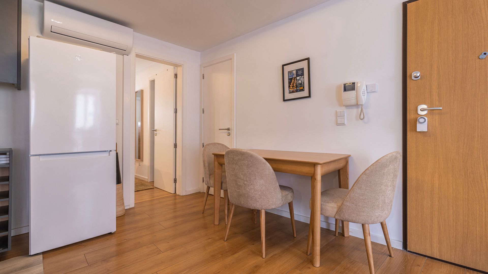 A compact kitchen with a refrigerator, a wooden dining table, and two chairs. There is a door leading to another room and a wall-mounted phone.