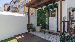 Un pequeño patio al aire libre con terraza de madera, dos sillas de metal, una puerta blanca y una pared verde con flores rojas.