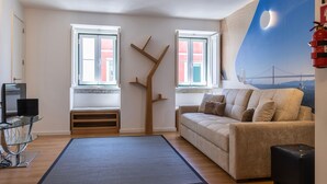 Un salón con sofá, una mesa de centro de vidrio, un banco de madera y dos ventanas con vistas a edificios.