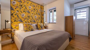 Un dormitorio con cabecera de flores, cama con ropa de cama blanca y gris, mesita de noche con lámpara y armario de madera.