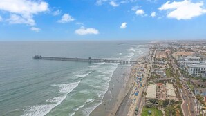 Un paisaje urbano costero con un muelle largo que se adentra en el mar, una playa con gente y sombrillas, y edificios a lo largo de la orilla.