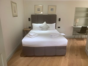 A neatly made bed with pillows and folded towels, a bedside table, a chair, and a desk with a lamp. - Kensington Lodge - mins from Natural History Museum, on award-winning Gdn Sq  (London)
