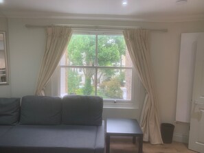 A room with a grey sofa, a small table, a window with curtains, and a white door. - Kensington Lodge - mins from Natural History Museum, on award-winning Gdn Sq  (London)